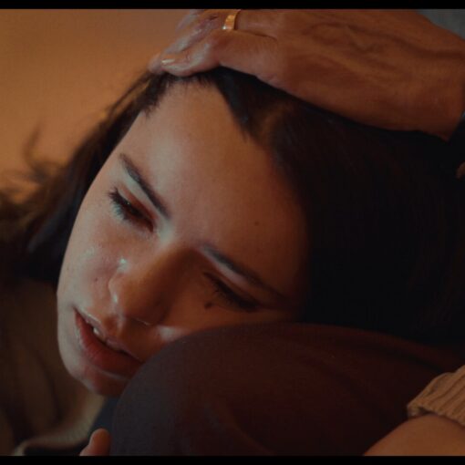 Delia, una joven con el rostro visiblemente emocionado, apoya la cabeza en el regazo de alguien mientras una mano adulta le acaricia con ternura el cabello. La escena está iluminada con luz cálida y transmite un momento íntimo de consuelo y vulnerabilidad emocional.