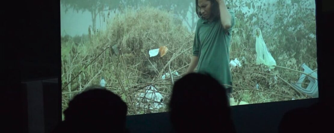 Audiencia viendo una película proyectada en una pantalla, mostrando a una persona con camisa verde entre escombros naturales.