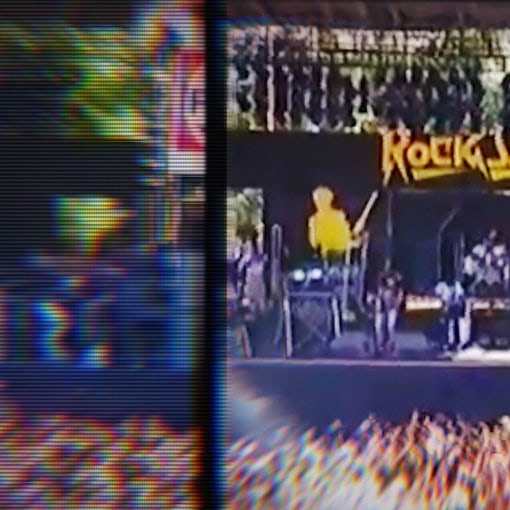 Una vista de un escenario de concierto al aire libre con una banda tocando y una multitud de personas. El escenario tiene el nombre "ROCK SANDER" en la parte superior.