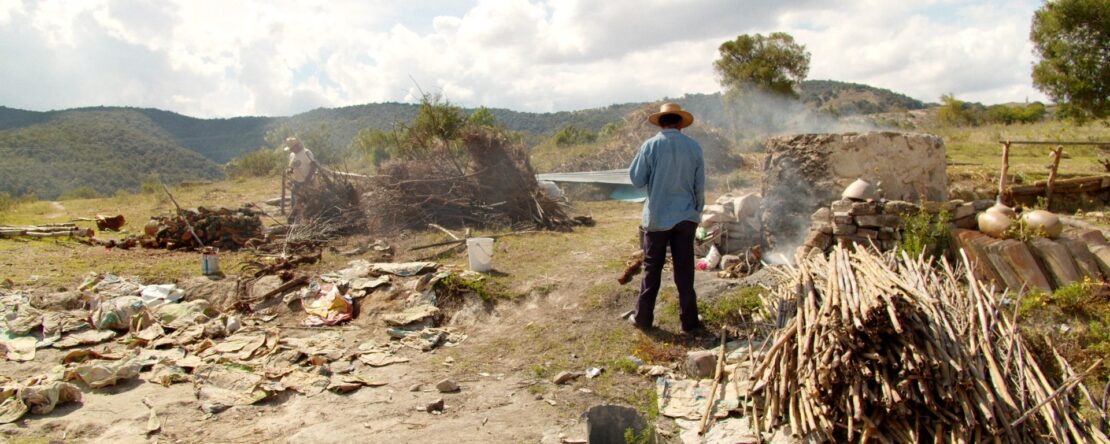Un hombre con sombrero y camisa azul de espaldas, observando un paisaje campestre con estructuras rústicas y montones de leña. Se aprecian columnas de humo.