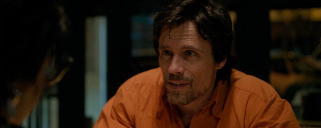 Fotograma de A una bala (2024) en el que un hombre con camisa naranja conversa en un interior con iluminación tenue.