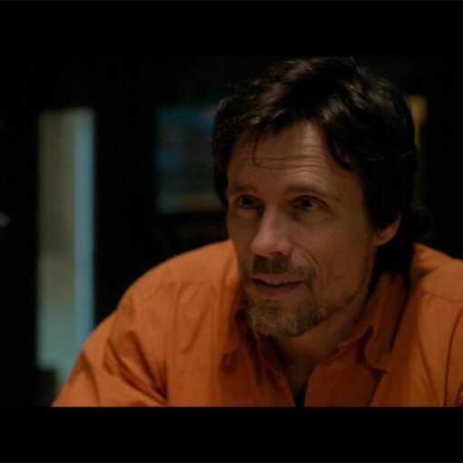 Fotograma de A una bala (2024) en el que un hombre con camisa naranja conversa en un interior con iluminación tenue.