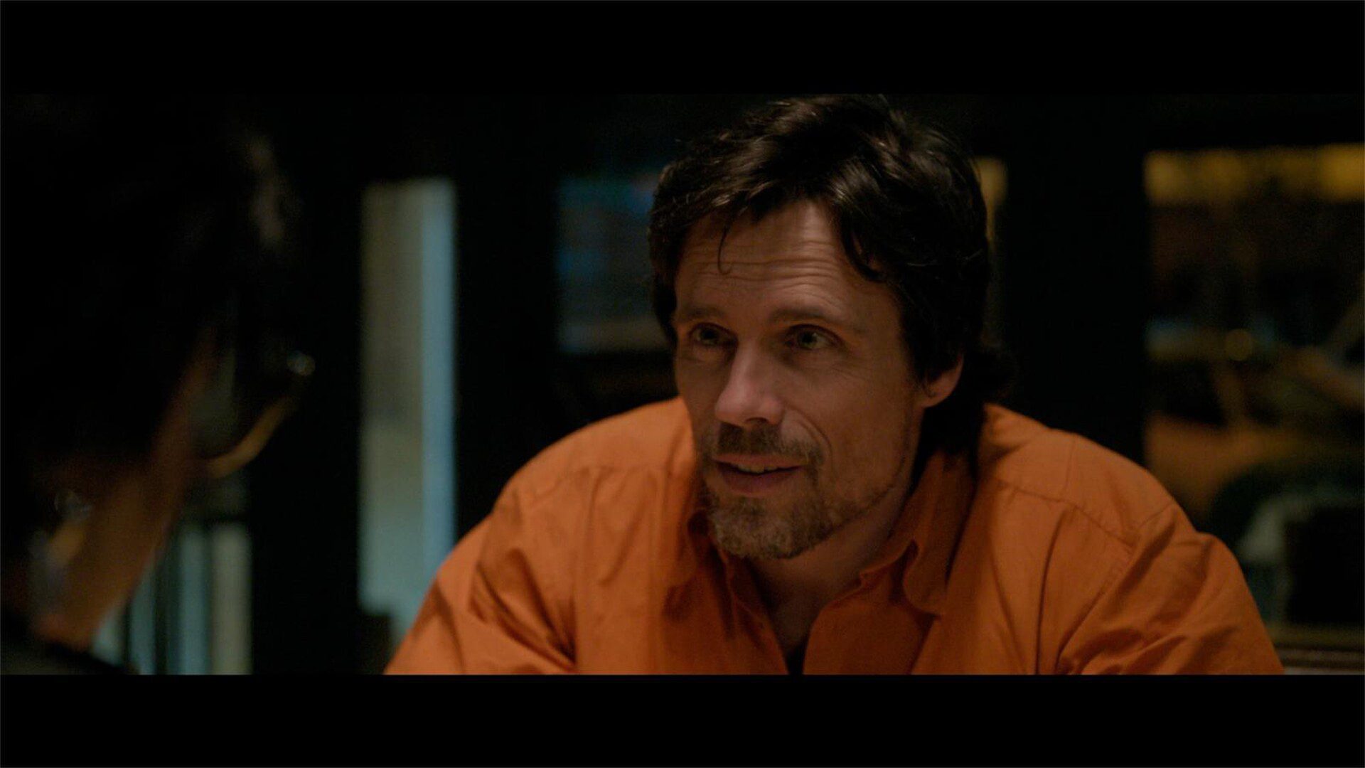Fotograma de A una bala (2024) en el que un hombre con camisa naranja conversa en un interior con iluminación tenue.