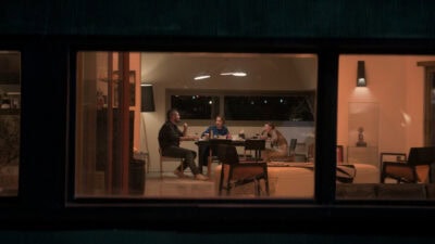 Una vista a través de una ventana de tres personas sentadas alrededor de una mesa en el interior de una casa, con luces cálidas y el exterior oscuro.