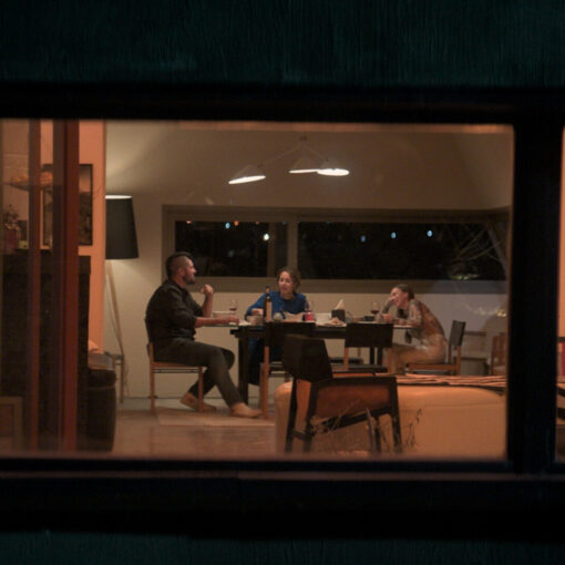 Una vista a través de una ventana de tres personas sentadas alrededor de una mesa en el interior de una casa, con luces cálidas y el exterior oscuro.