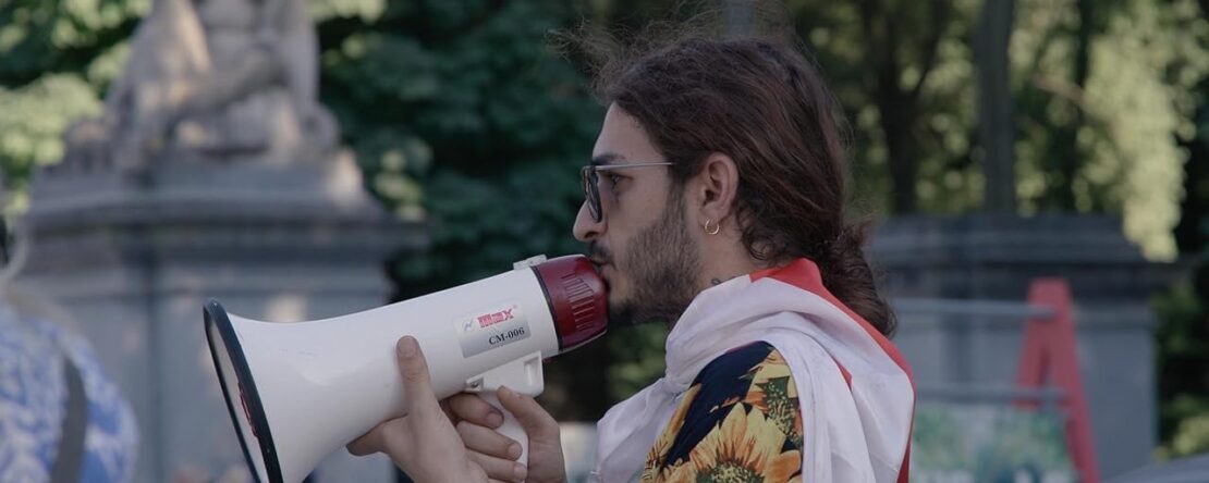 Activista LGBTQ+ de perfil usando un megáfono durante una manifestación. Lleva gafas, pendiente y una camisa colorida con girasoles. Escena del documental El mentiroso del orgullo.