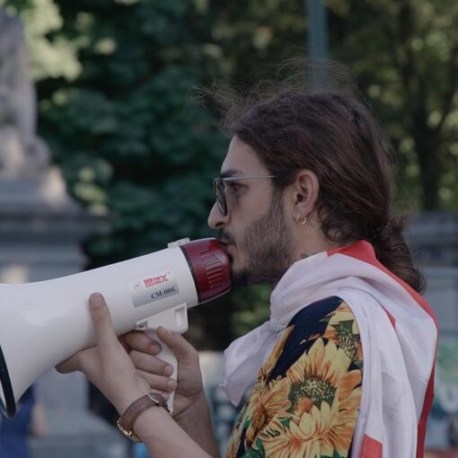 Activista LGBTQ+ de perfil usando un megáfono durante una manifestación. Lleva gafas, pendiente y una camisa colorida con girasoles. Escena del documental El mentiroso del orgullo.