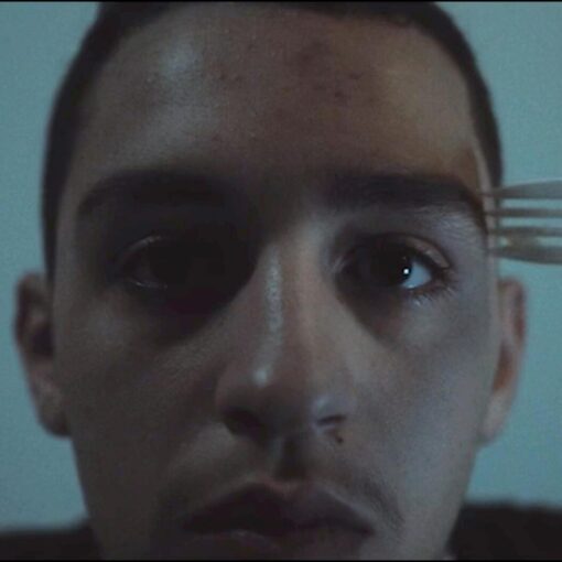 Primer plano de un hombre con una expresión de temor o incomodidad, mientras una mano sostiene un tenedor de plástico blanco cerca de su frente. La iluminación es tenue y fría.