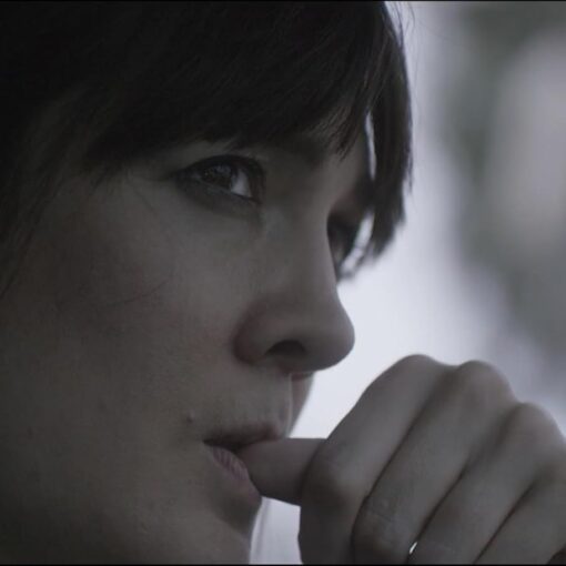Fotograma en primer plano del cortometraje La Pura Vida. Se muestra el rostro de una mujer joven con expresión seria y mirada perdida, mientras se lleva la mano a la boca. La escena transmite introspección y angustia emocional, enmarcada por una iluminación suave y desenfocada.