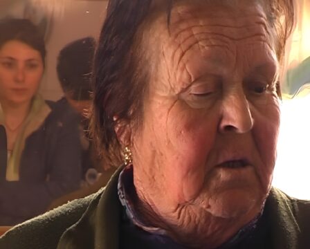 Primer plano del rostro arrugado de una mujer mayor con un pendiente dorado. Al fondo, se ven dos personas reflejadas en un cristal.