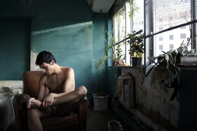 Un joven marroquí, con el torso desnudo, está sentado pensativo en un sillón dentro de una habitación deteriorada. La luz entra por la ventana, iluminando varias plantas y creando un ambiente melancólico.