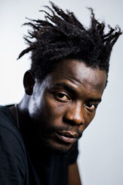 Fotografía del director haitiano Pierre Michael Jean, hombre de color con camiseta negra y peinado afro. Mirada a cámara por lo que esta realizada en un estudio.