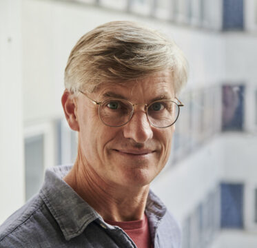 Fotografía retrato del director Rich Minnich posando con una camisa vaquera fina de color gris, camiseta interior de color roja y lleva unas gafas de actualidad de montura fina y redondeadas.