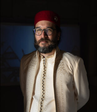 Hombre árabe posando con las manos agarradas a su espalda. Viste ropa tradicional árabe de color beige y gorro de color rojo.