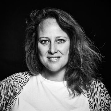 Fotografía de la directora Barbara de Baare en blanco y negro. Mirando a cámara y sonriendo con melena pasados los hombros, piel blanca y con camiseta básica blanca y una especia de cárdigan estampado.