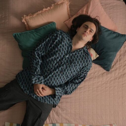 Fotograma de la película Temas Propios. Un joven con cabello largo y camisa azul oscura está acostado boca arriba sobre una cama con colcha rosa. Alrededor hay cojines de colores, una alfombra y una mesa de noche con libros y objetos personales. Su expresión transmite introspección.