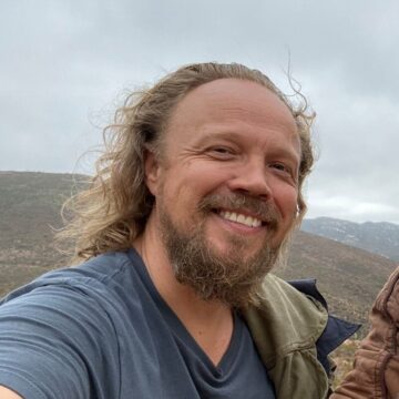 Fotografía selfie del director del largometraje Pégame Dylan Verrechia con una camiseta azul y de fondo el monte y nubes.