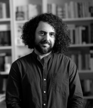 Fotografía en blanco y negro de Baran Gunduzalp, director del largometraje Rosinante, posando con una camisa de color oscuro. Tiene el cabello negro, rizado tipo crespo y media melena.
