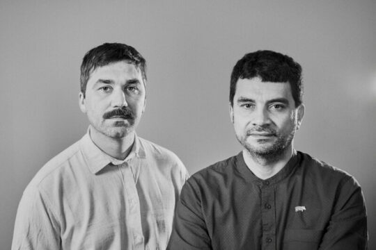 Fotografía en blanco y negro de los dos directores del documental Sobre hipopótamos felices y pavos reales tristes. Por la izquerda, Johannes föster, viste camisa blanca, corte de pelo corto y bigote con rasgos alemanes y a la derecha, Elkin Calderón, vistiendo camisa oscura, corte de pelo corto y barba corta y arreglada con rasgos latinos.