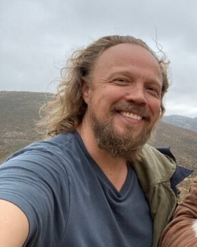 El director de Tierra Kumeyaay Dylan Verrechia, de cabello largo y barba sonriendo al aire libre, con colinas al fondo en un día nublado.