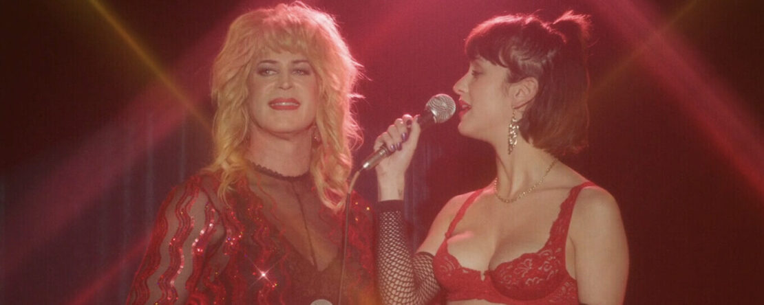 Fotograma de la película Cuatro Estrellas donde aparecen una mujer trans al lado de otra en un escenario, mientras sostienen dos micrófonos y llevan a cabo una actuación.