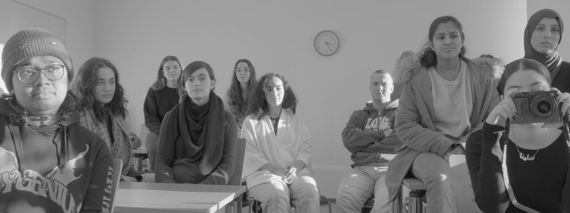 Grupo diverso de personas jóvenes sentadas en un aula, mirando directamente a la cámara; una persona en primer plano sostiene una cámara fotográfica.