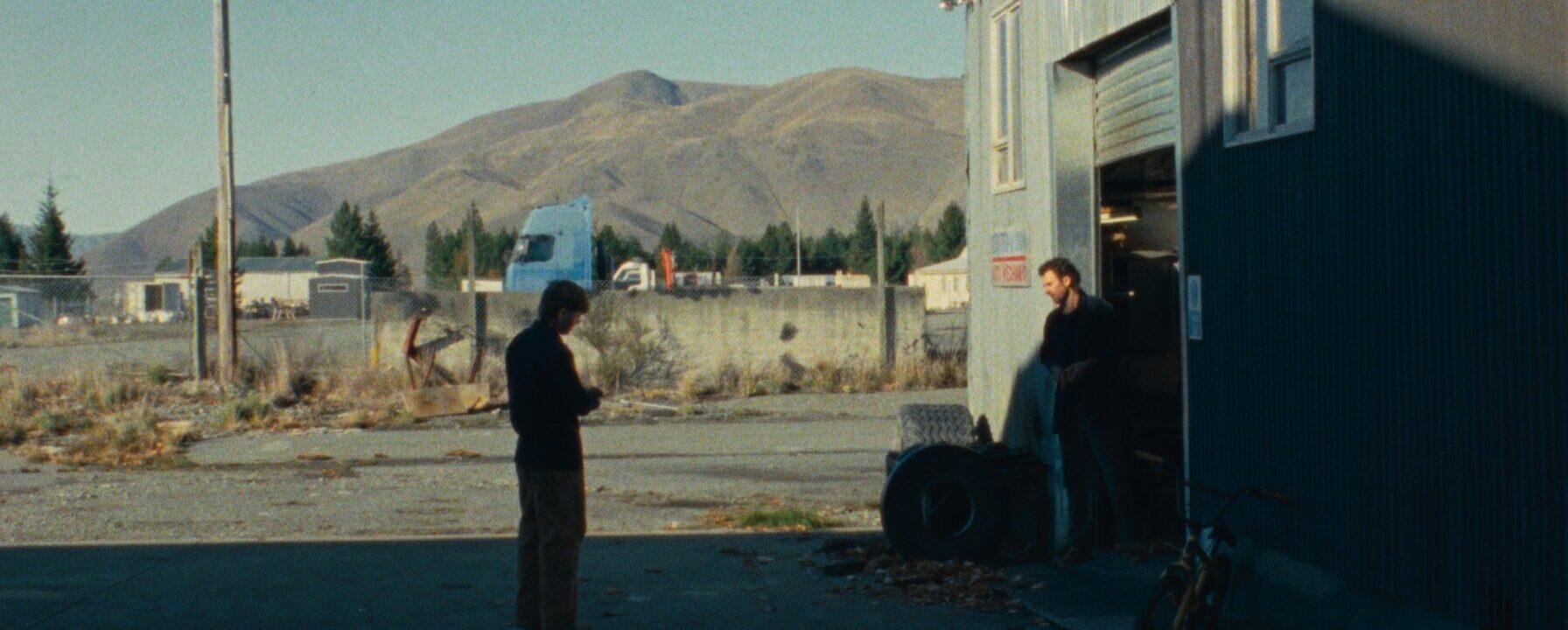 Dos hombres de pie frente a un taller industrial en una zona rural, con montañas al fondo y una bicicleta apoyada en la pared.