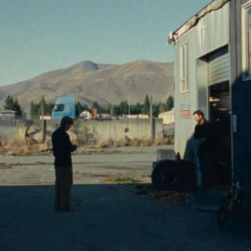 Dos hombres de pie frente a un taller industrial en una zona rural, con montañas al fondo y una bicicleta apoyada en la pared.
