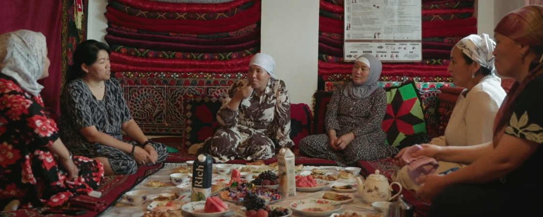 Mujeres sentadas en el suelo comparten comida y conversan en una habitación decorada con alfombras tradicionales.