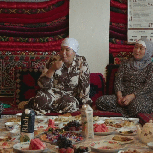Mujeres sentadas en el suelo comparten comida y conversan en una habitación decorada con alfombras tradicionales.