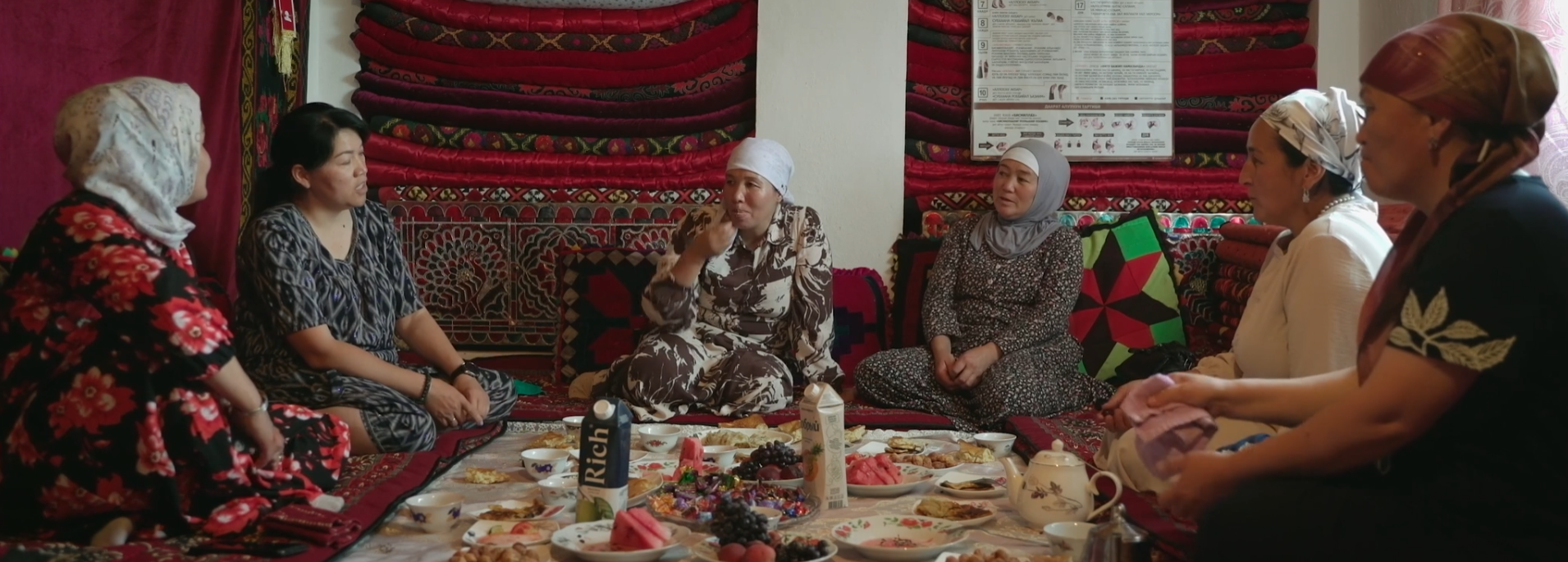 Mujeres sentadas en el suelo comparten comida y conversan en una habitación decorada con alfombras tradicionales.