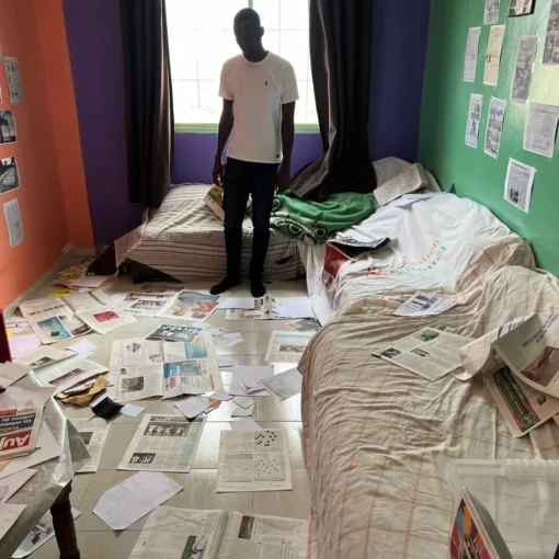 Fotograma de 2002 Batalla contra el olvido en el que aparece un hombre de pie en una habitación desordenada con el suelo y las paredes llenas de páginas de periódicos.