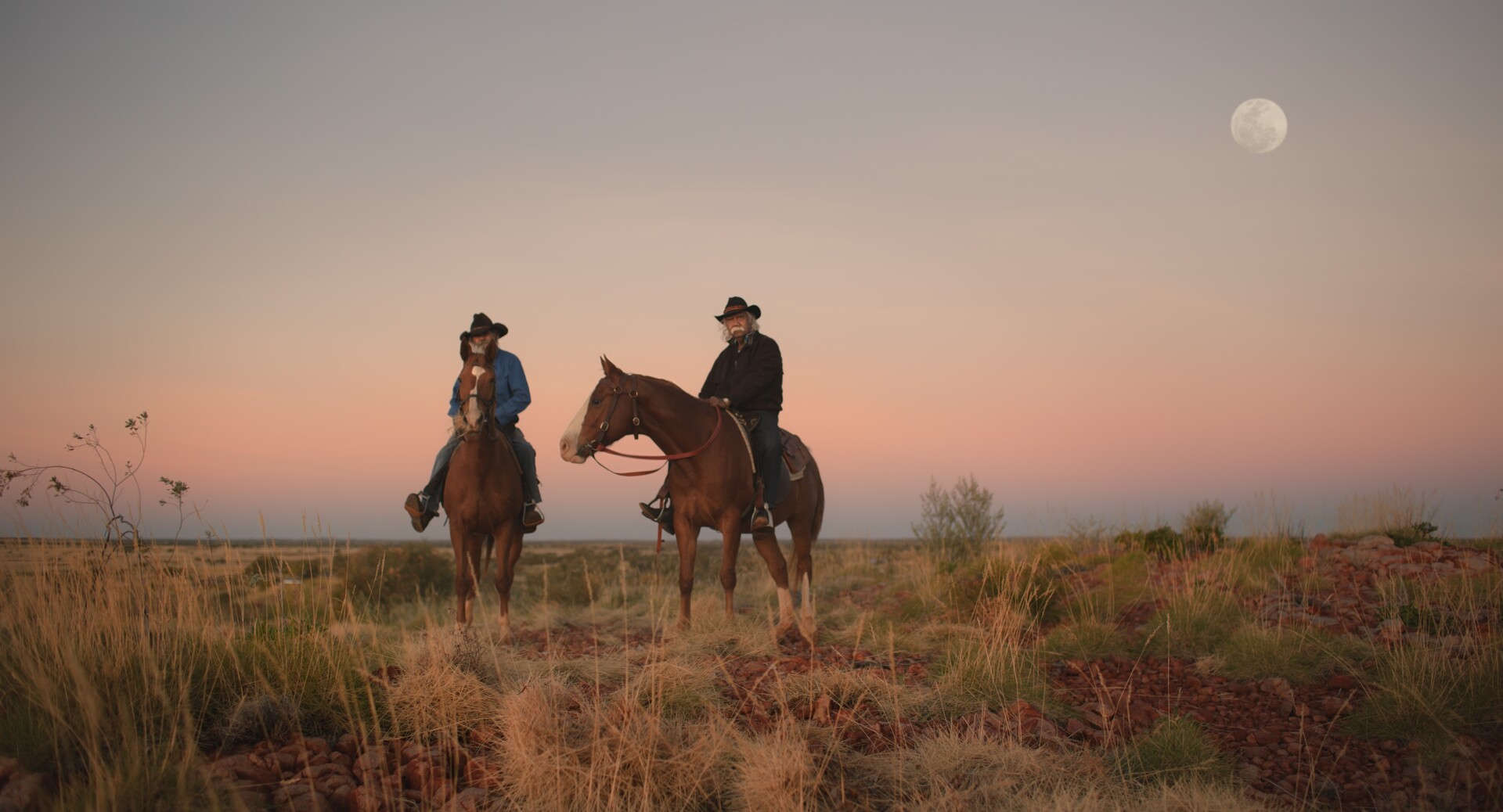 Fotograma de Yurlu | País dos hombres vestidos de vaqueros sobre caballos en un paisaje desértico. En el cielo, está la luna.