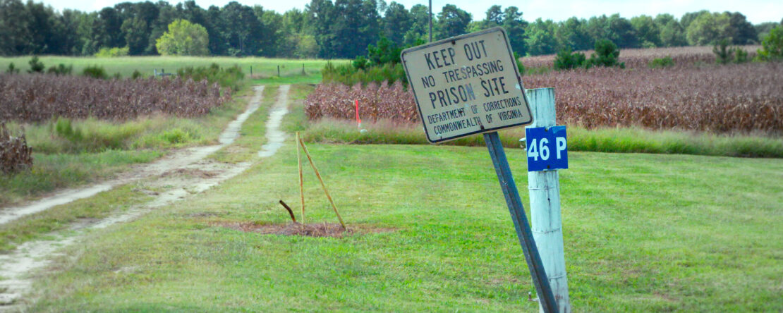 Cartel inclinado que advierte “Keep Out – Prison Site” junto a un camino rural que atraviesa un campo abierto.