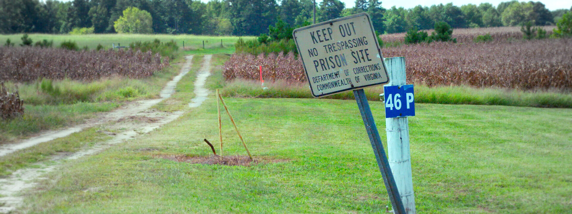 Cartel inclinado que advierte “Keep Out – Prison Site” junto a un camino rural que atraviesa un campo abierto.