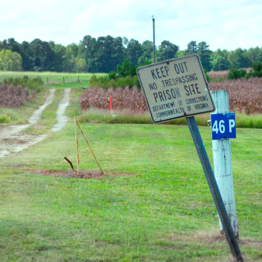 Cartel inclinado que advierte “Keep Out – Prison Site” junto a un camino rural que atraviesa un campo abierto.