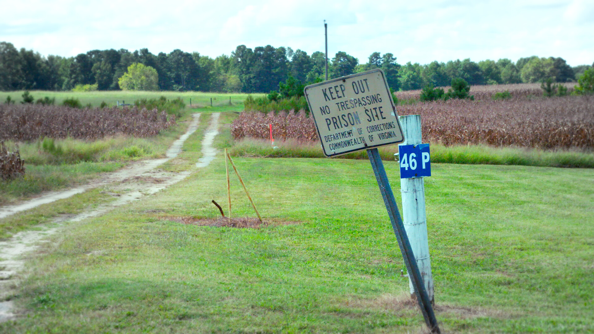Cartel inclinado que advierte “Keep Out – Prison Site” junto a un camino rural que atraviesa un campo abierto.