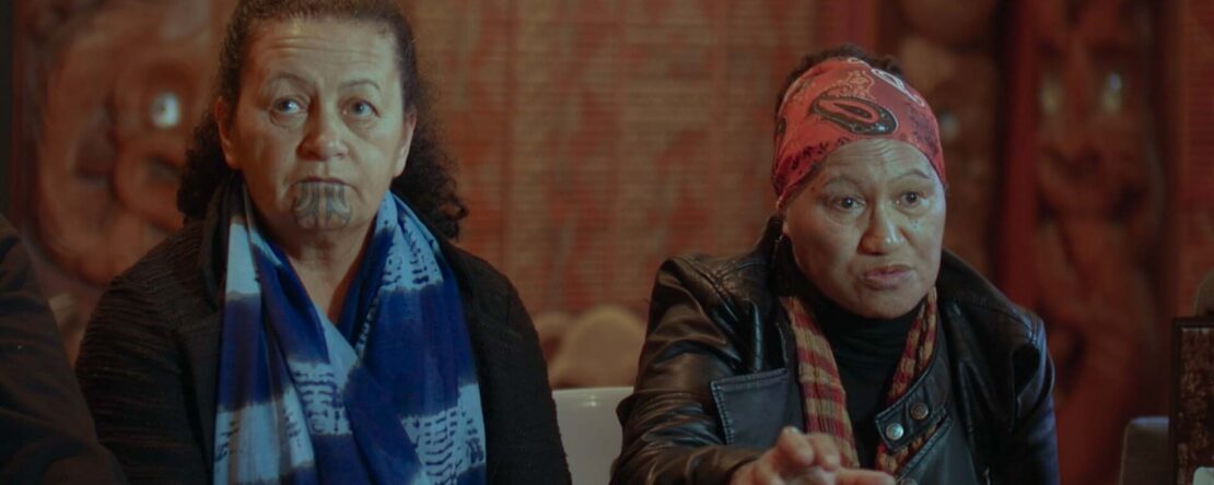 Dos mujeres maoríes conversan en una entrevista en interior; una de ellas lleva un tatuaje tradicional moko en la barbilla.