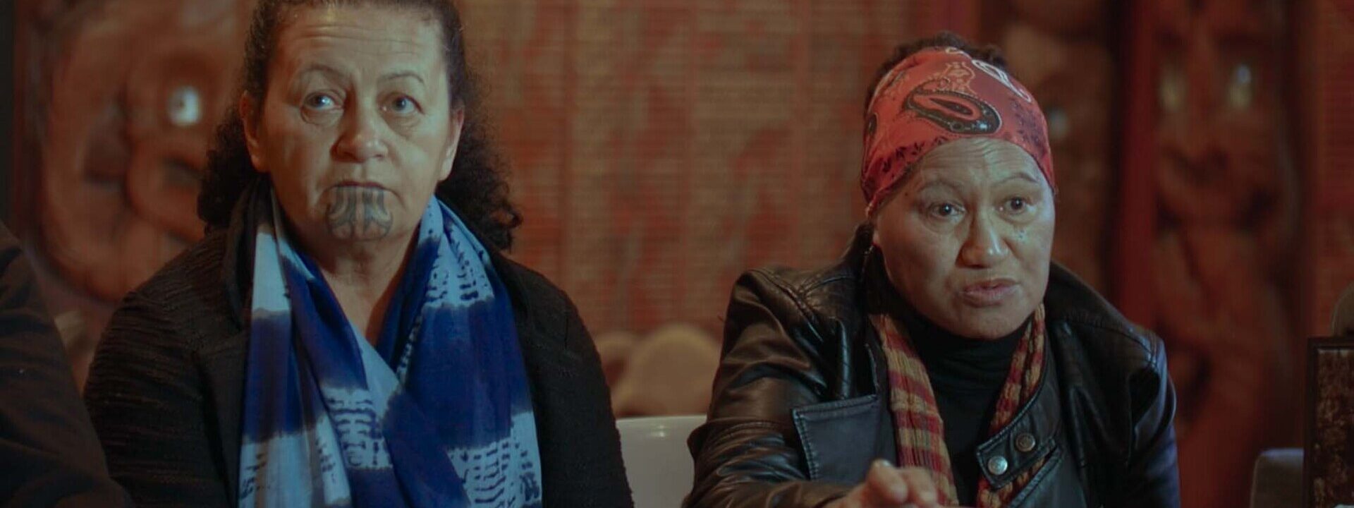 Dos mujeres maoríes conversan en una entrevista en interior; una de ellas lleva un tatuaje tradicional moko en la barbilla.