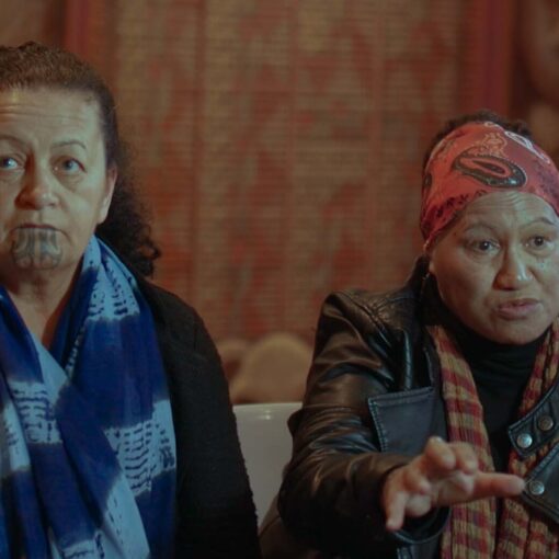 Dos mujeres maoríes conversan en una entrevista en interior; una de ellas lleva un tatuaje tradicional moko en la barbilla.