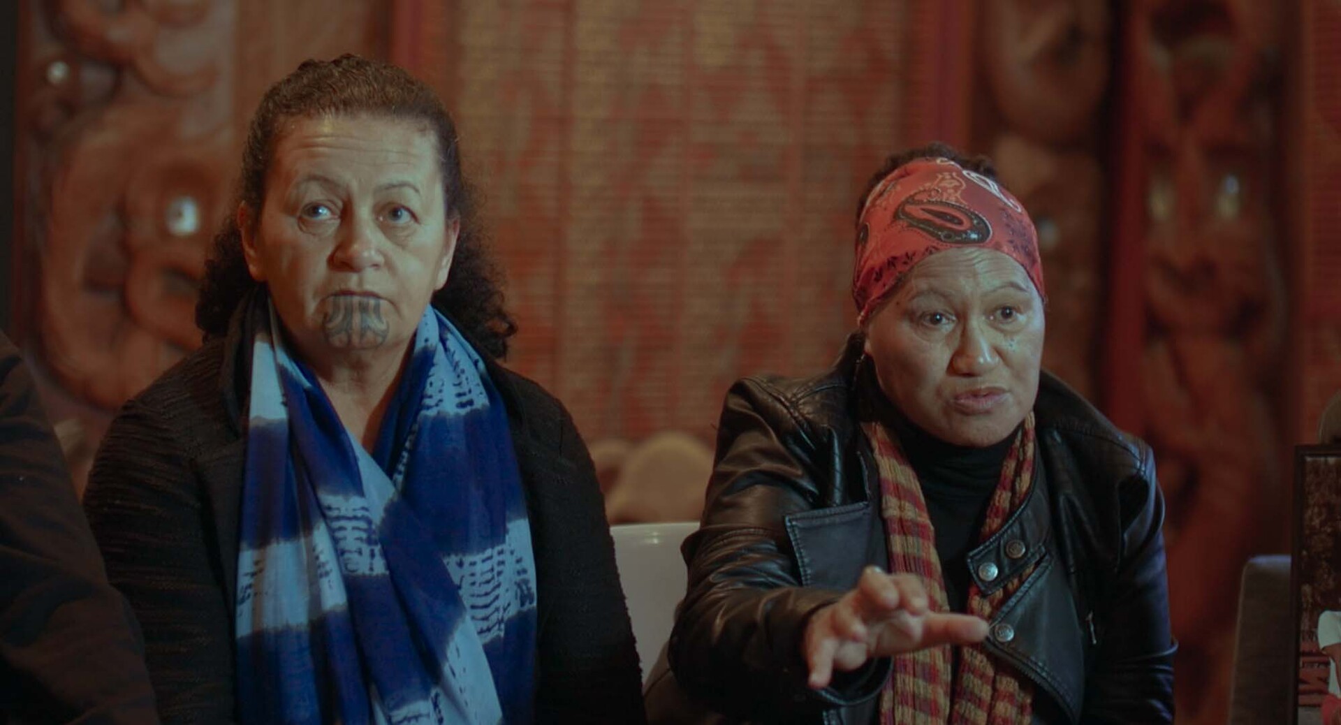 Dos mujeres maoríes conversan en una entrevista en interior; una de ellas lleva un tatuaje tradicional moko en la barbilla.